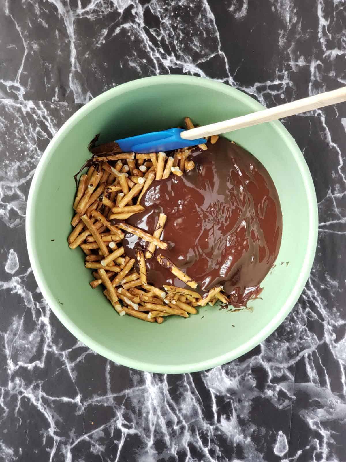 Mixing pretzel sticks and melted chocolate in green bowl to make easy clusters.