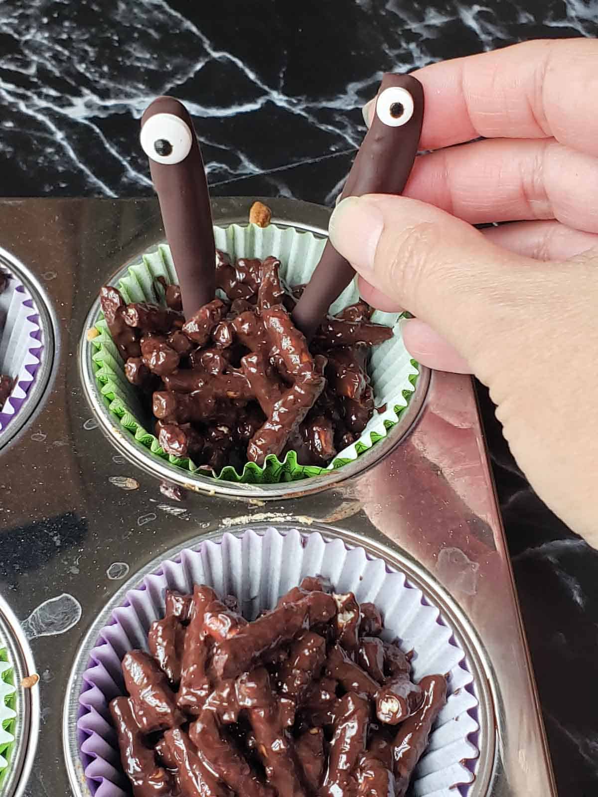 Hand placing chocolate covered pretzel sticks with candy eyes into chocolate clusters.