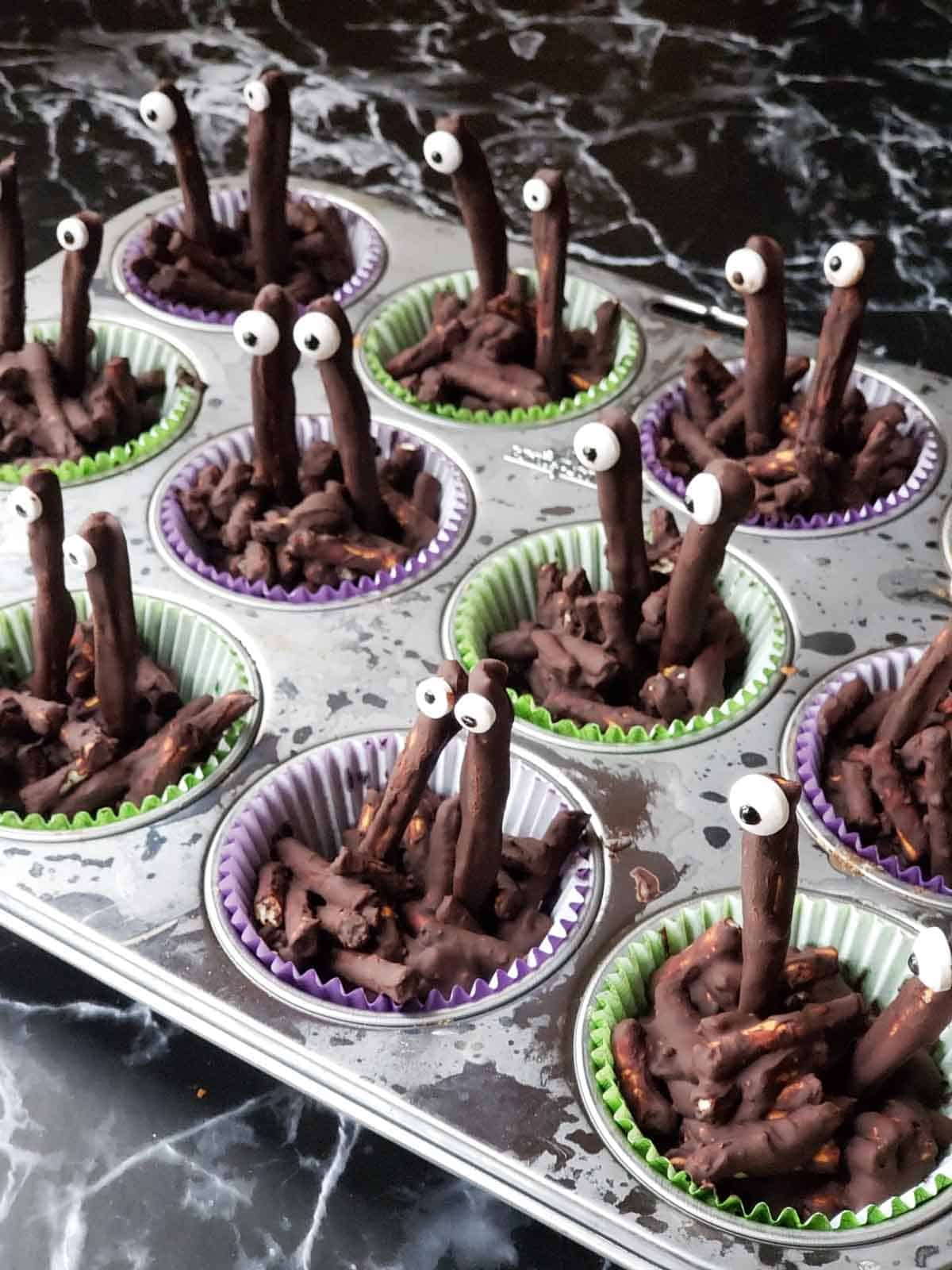 Close-up of chocolate snack clusters with candy eyes in orange and purple muffin liners.