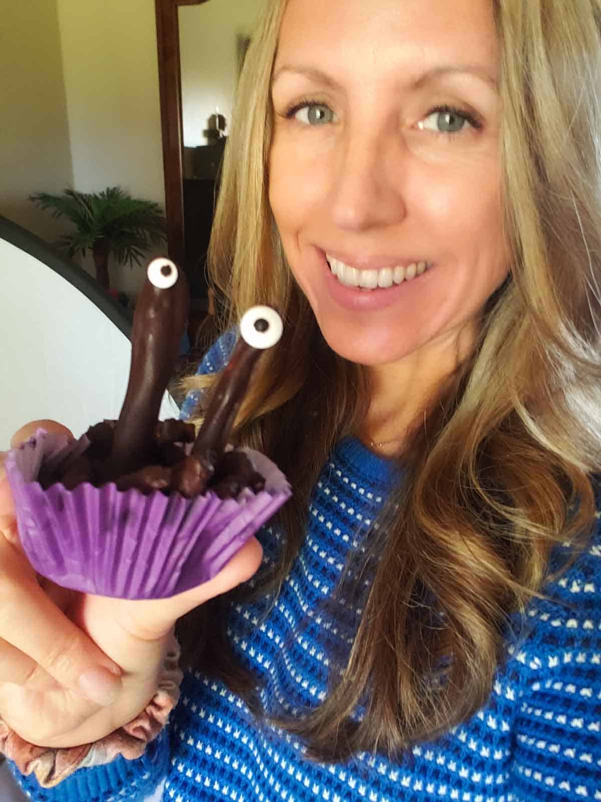 Smiling woman holding chocolate pretzel monster in purple cupcake liner for Halloween treat.