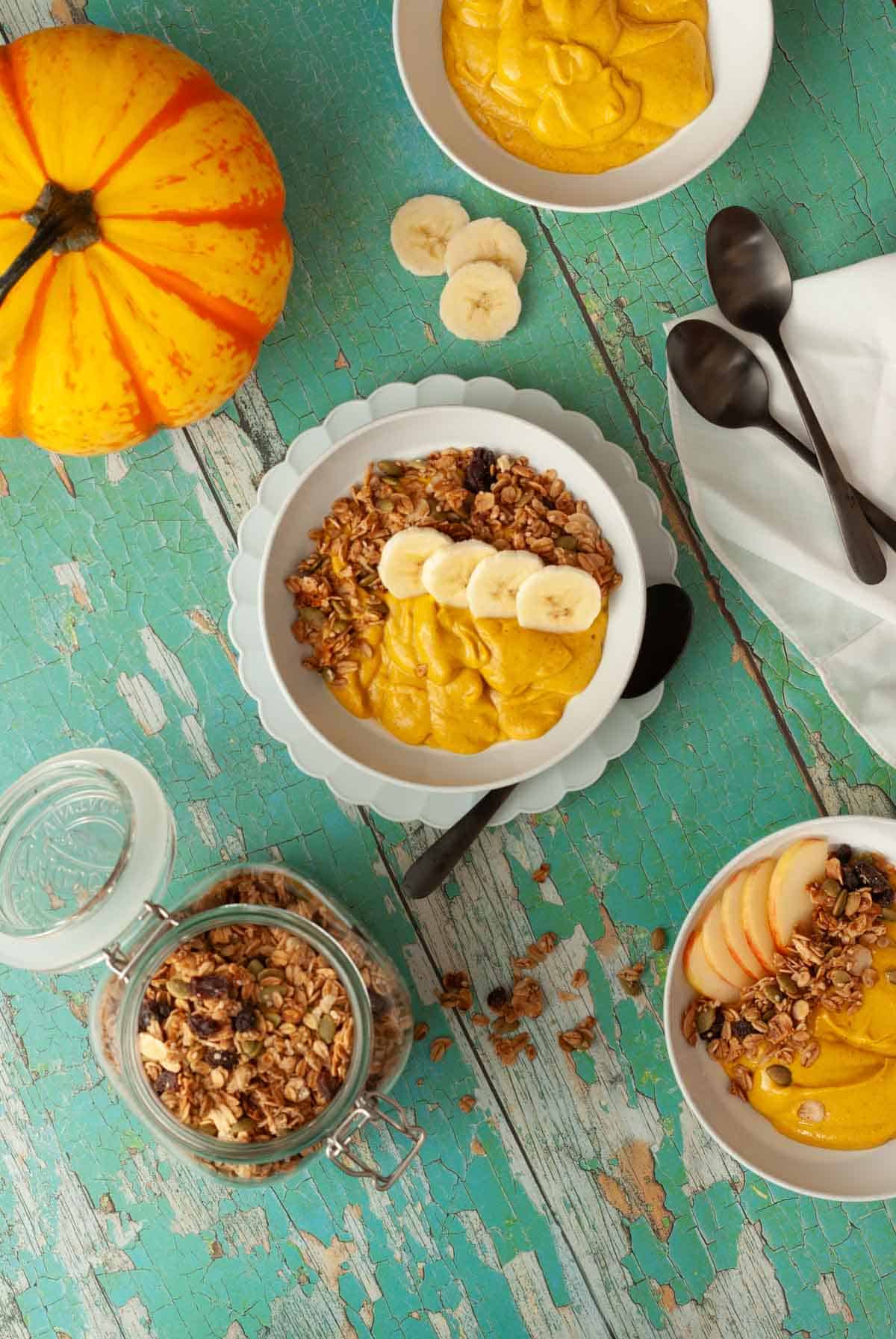 Overhead of vegan pumpkin breakfast bowls topped with granola and banana on a rustic turquoise table.