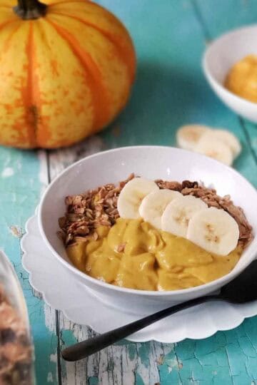 Creamy vegan pumpkin breakfast bowl with granola and banana slices served beside a bright orange pumpkin.
