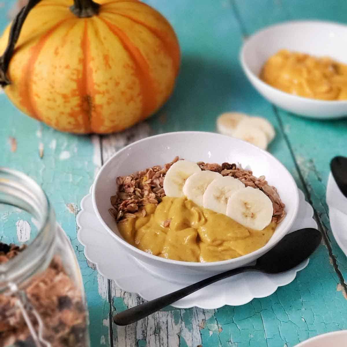 Creamy vegan pumpkin breakfast bowl with granola and banana slices served beside a bright orange pumpkin.