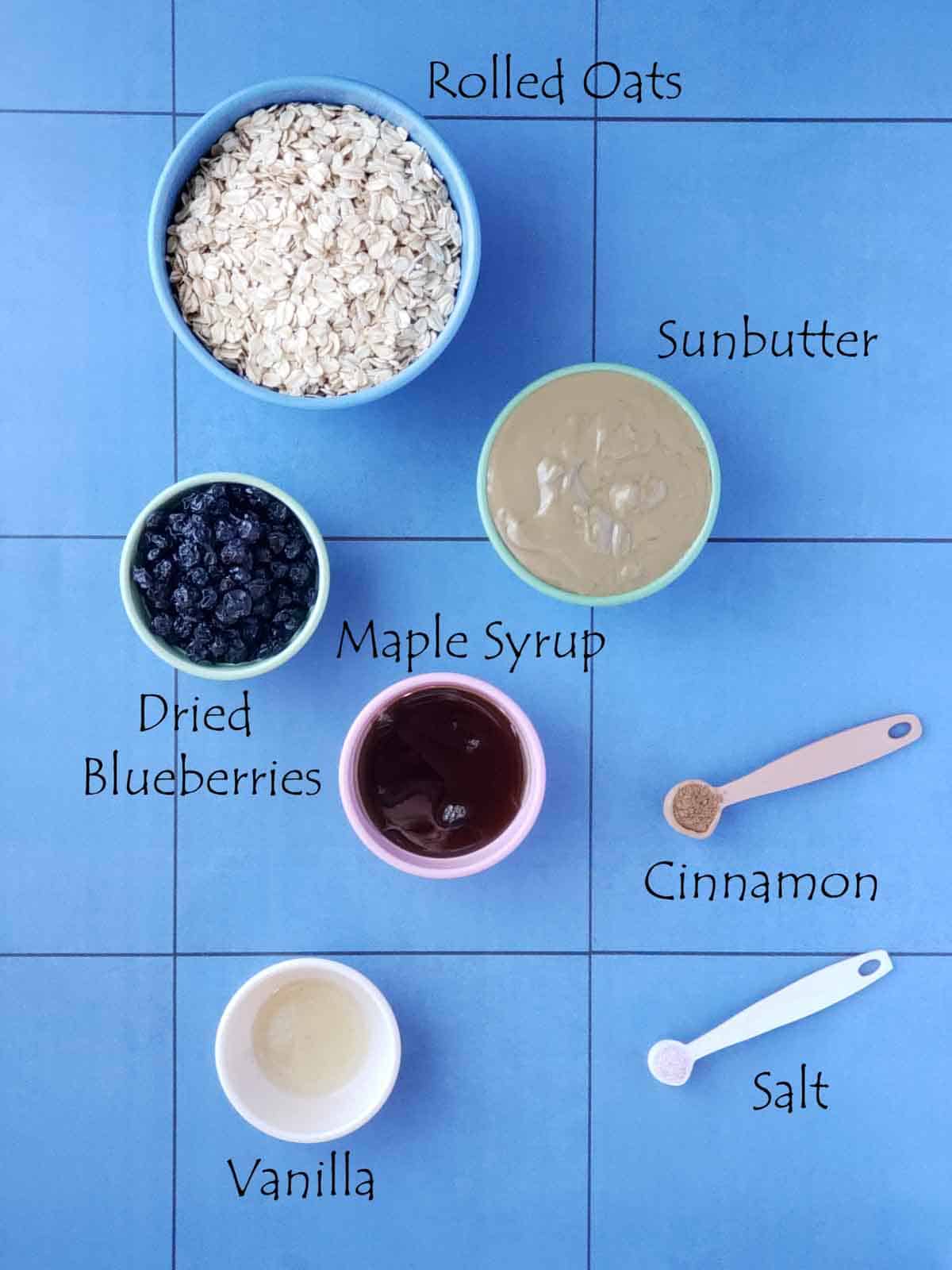 Overhead view of rolled oats, sunbutter, maple syrup, dried blueberries, vanilla, cinnamon, and salt measured out in bowls on a blue background.