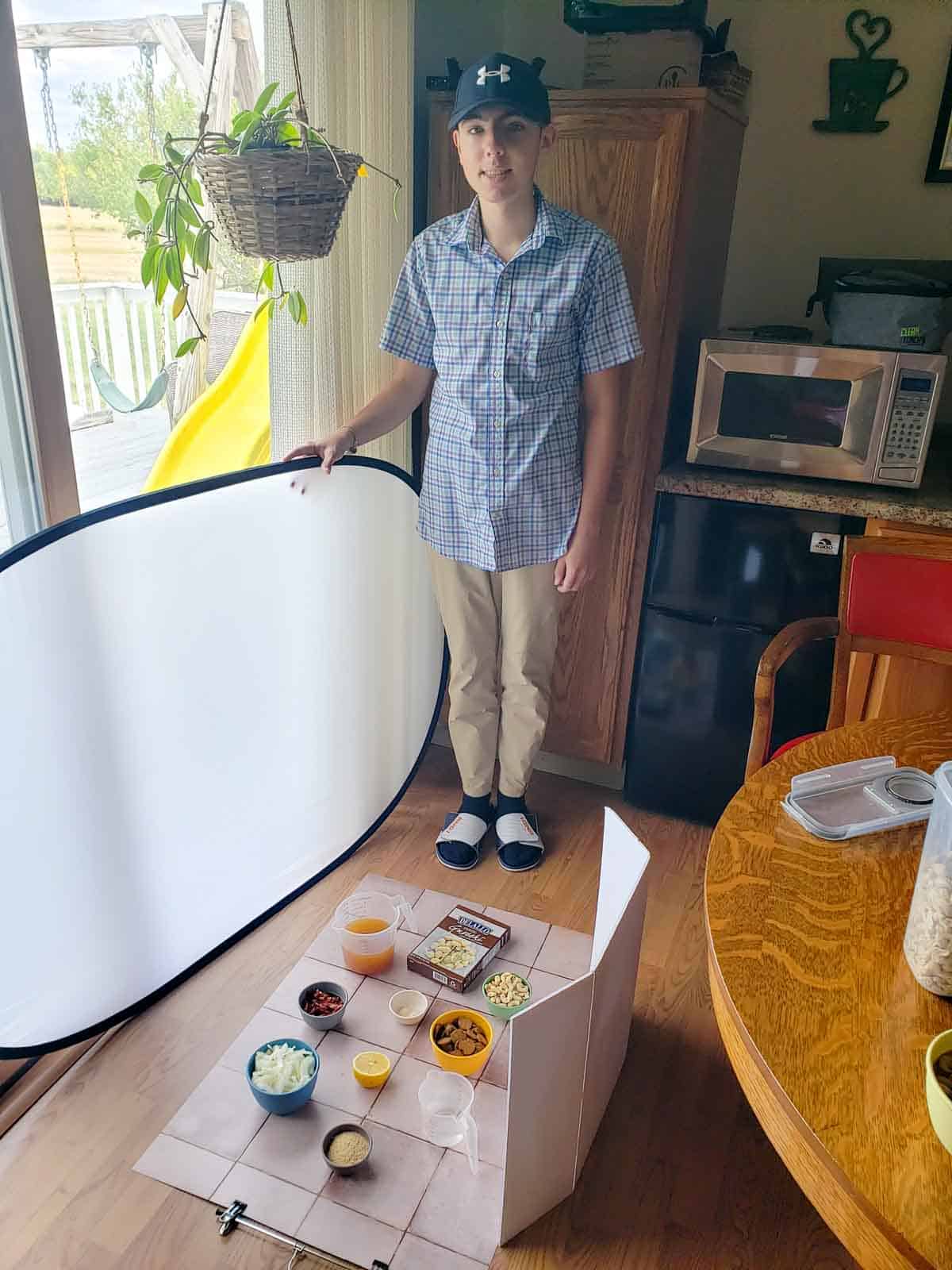 A teen boy standing beside a window with photo props and ingredients laid out on a tile board for a recipe shoot.