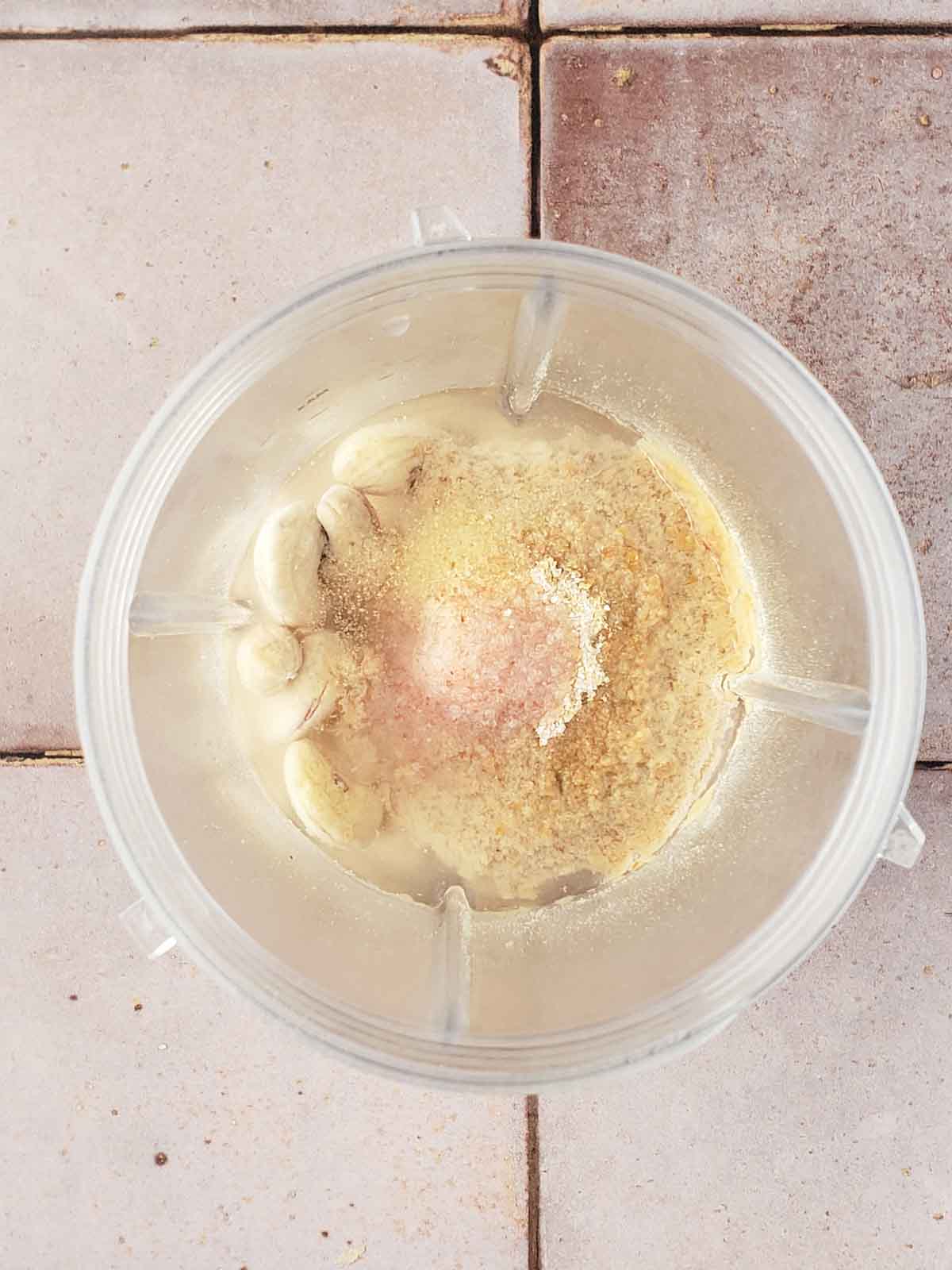 Cashews, nutritional yeast, salt, water, and garlic granules in a blender cup ready to blend for creamy vegan cashew sauce.