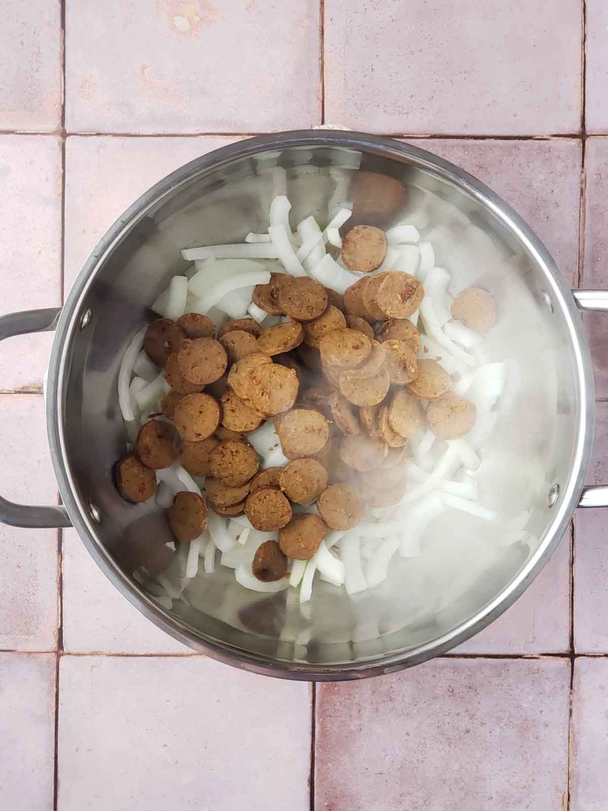 Sliced vegan Italian sausage and onions cooking in a large stainless steel pot with steam rising.