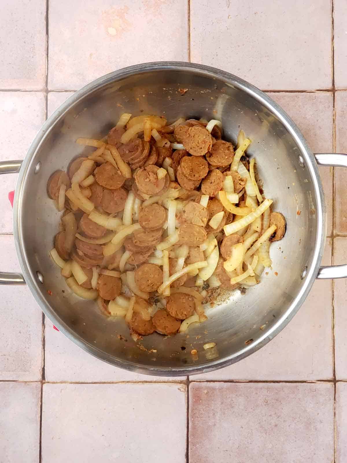 Vegan sausage and onions sautéed and lightly browned in a stainless steel pot after cooking for several minutes.