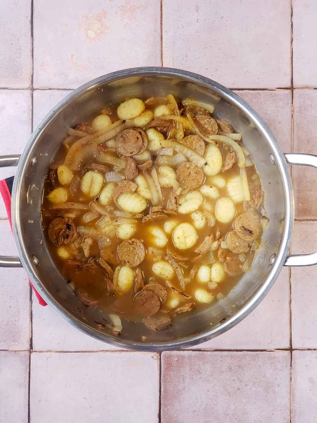 Gnocchi simmering in vegetable broth with onions and vegan sausage in a large pot.