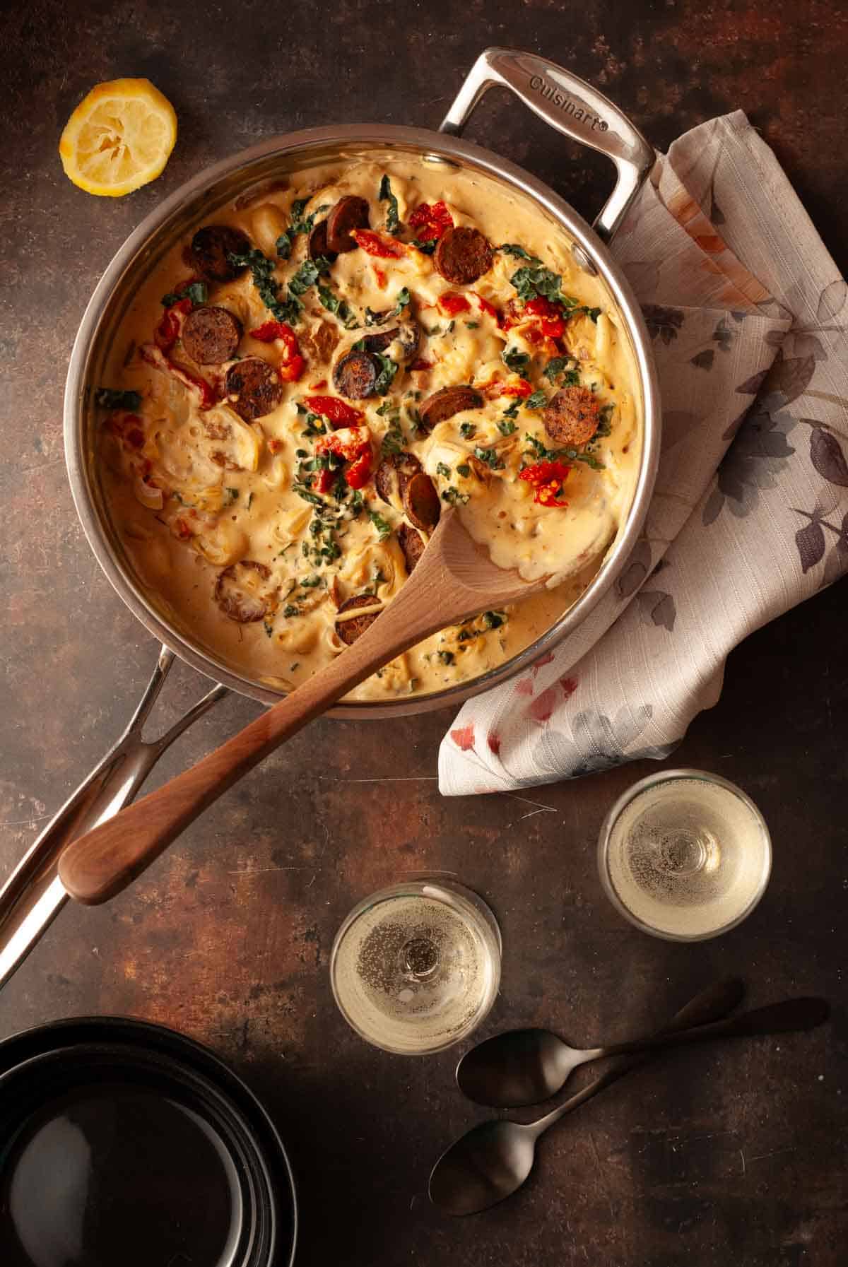 Overhead view of creamy vegan Tuscan gnocchi with kale, sun-dried tomatoes, and sausage in a stainless steel pan on a rustic table.
