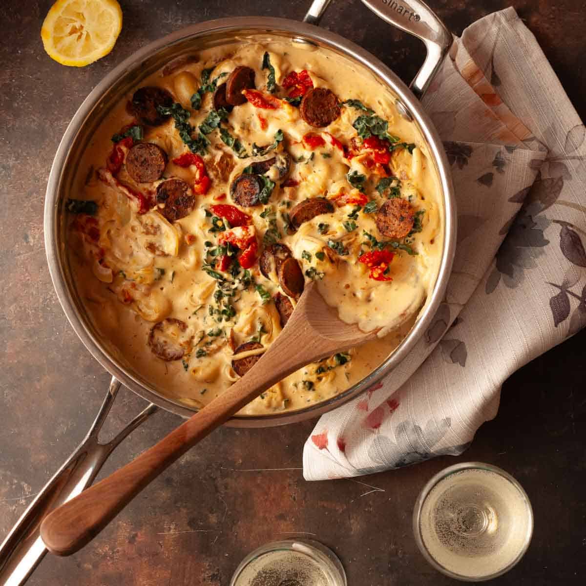 Close-up of creamy vegan Tuscan gnocchi with kale, sausage, and sun-dried tomatoes in a pan on a dark background.