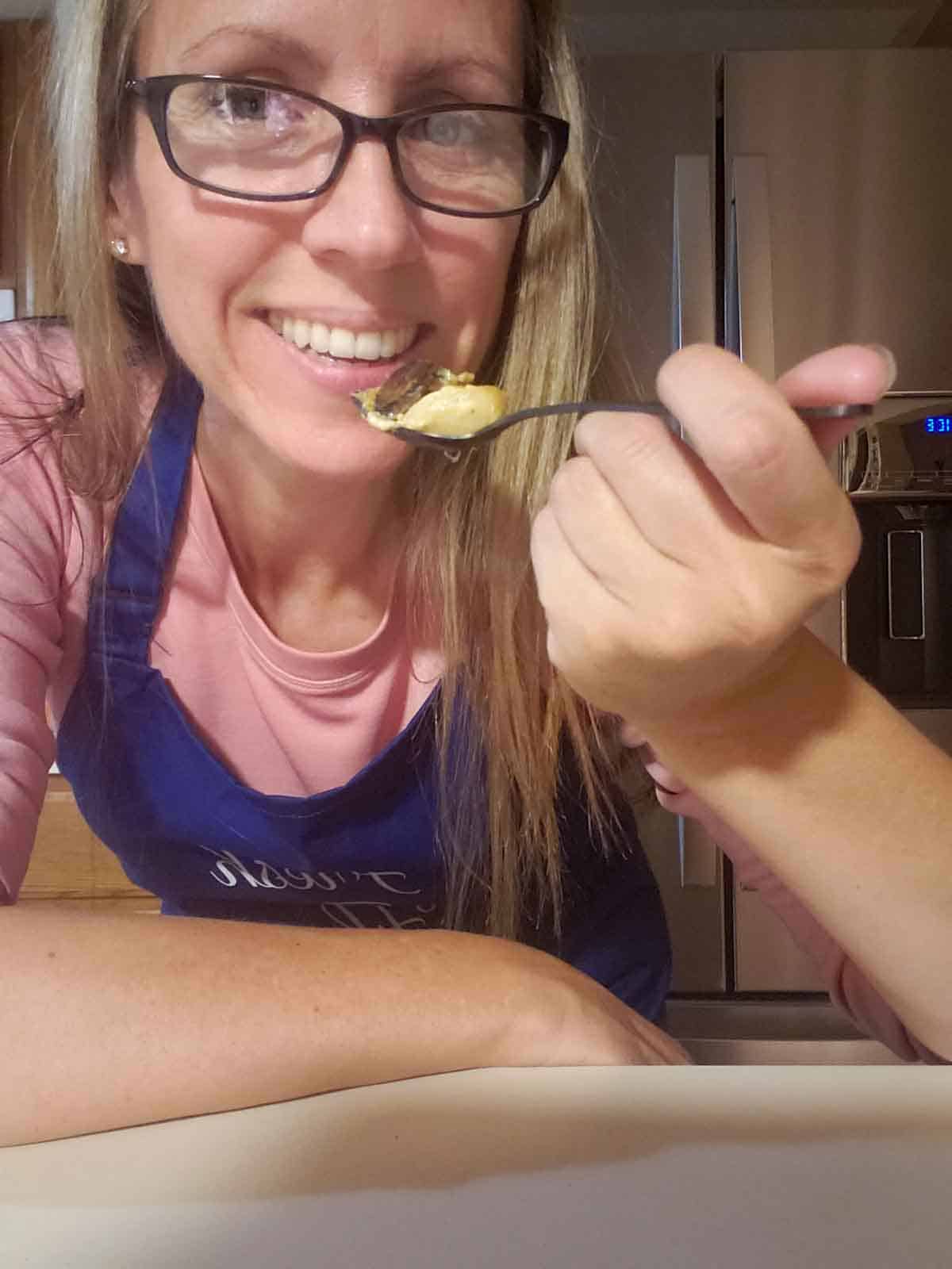 Woman smiling and holding a fork while wearing a pink shirt and blue apron.