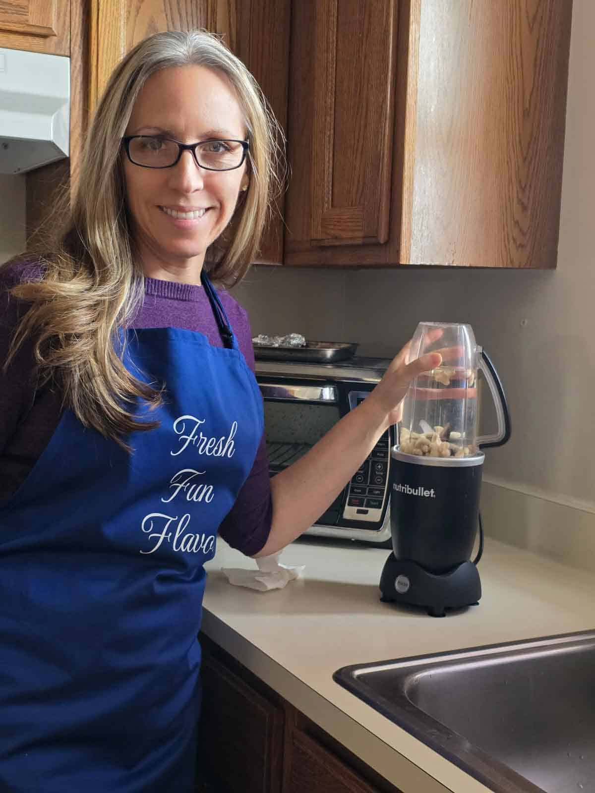 A woman wearing a Fresh Fun Flavor apron while blending cashews in a small countertop blender in her kitchen.
