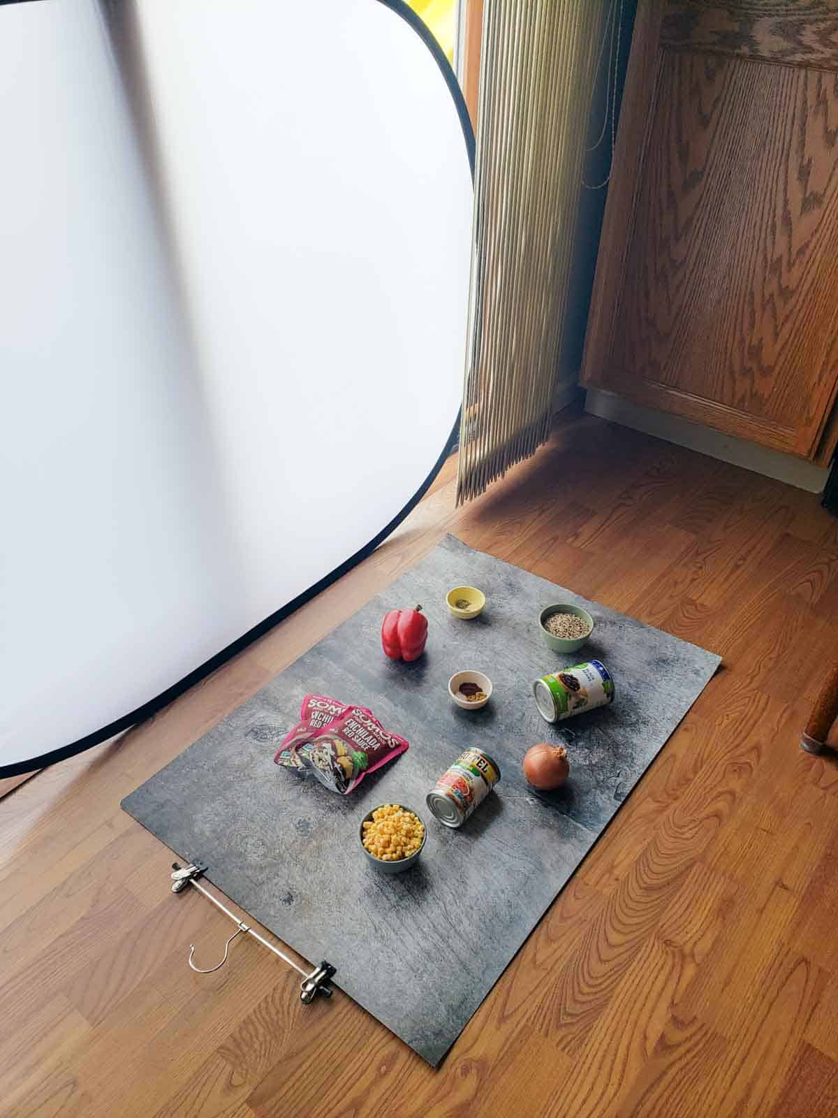 Behind-the-scenes photo setup for vegan enchilada bake ingredients on a gray surface, with cans, spices, and fresh vegetables ready for cooking.