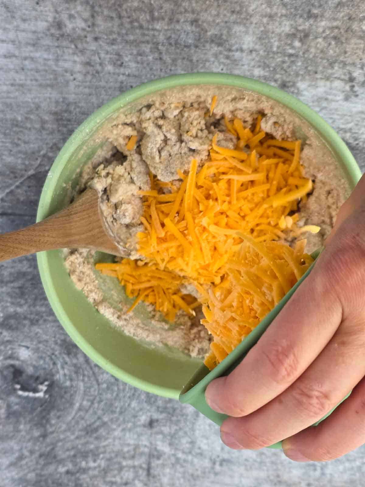 Hand adding shredded vegan cheese to the biscuit dough mixture.