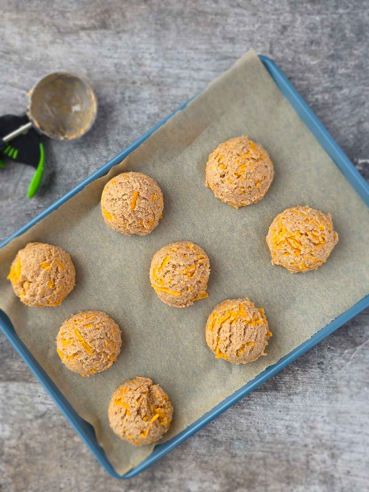 Scooped portions of dough for garlic cheese biscuits arranged on a parchment-lined baking sheet before baking.