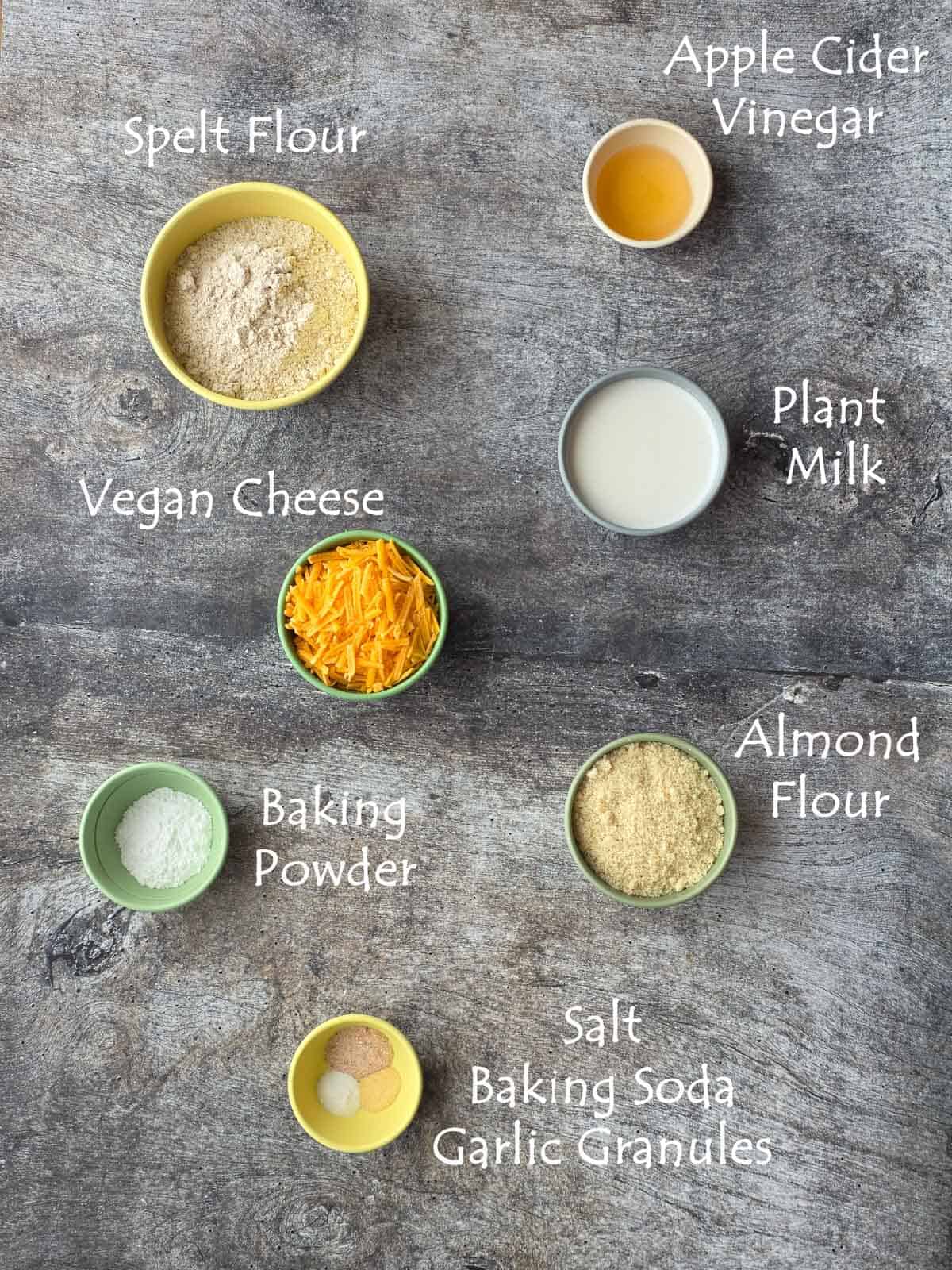 Flat lay of labeled ingredients in small bowls, including flours, plant milk, vinegar, baking powder, spices, and vegan cheese.
