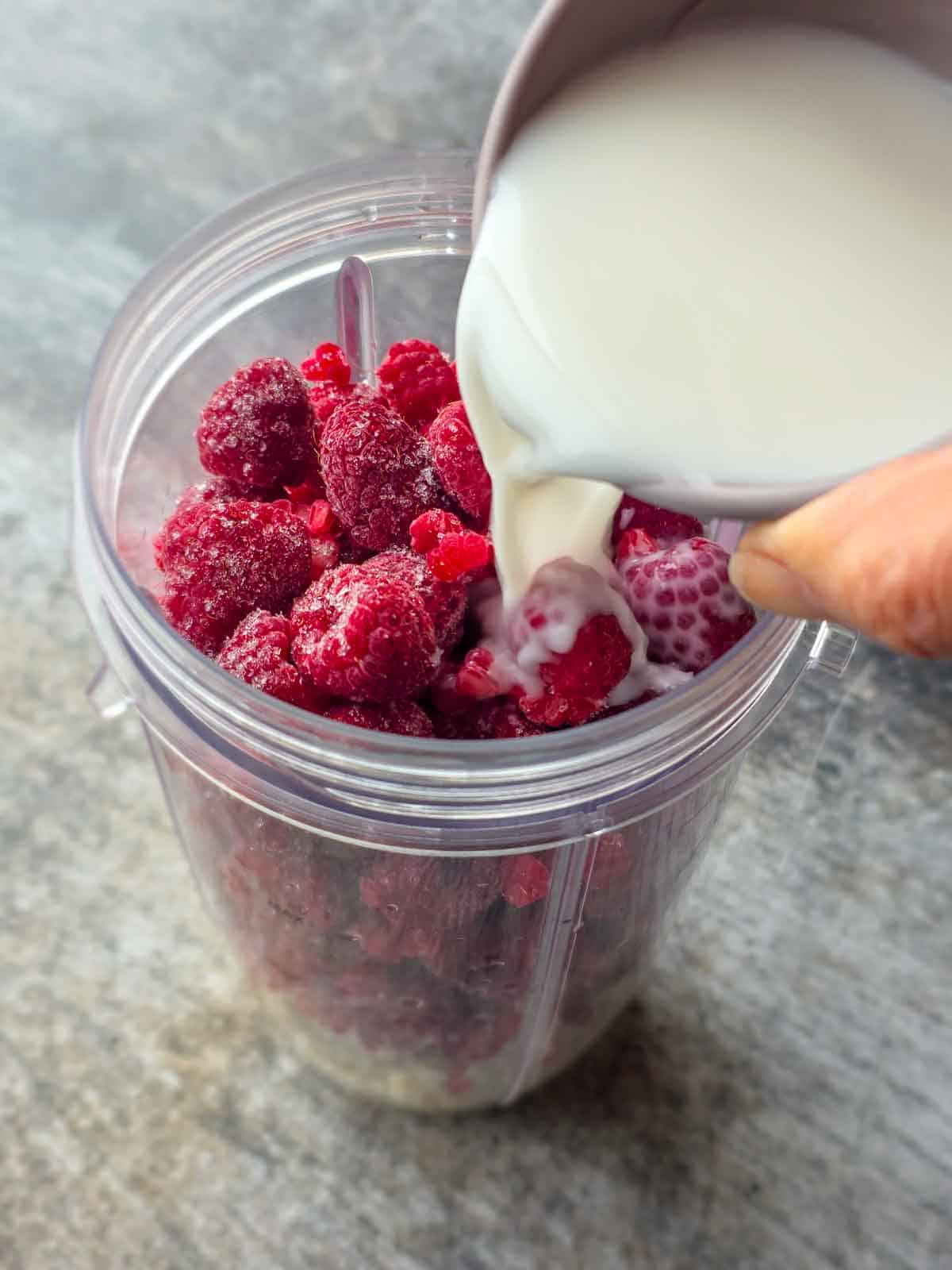 Plant milk being poured over frozen raspberries in a blender cup.