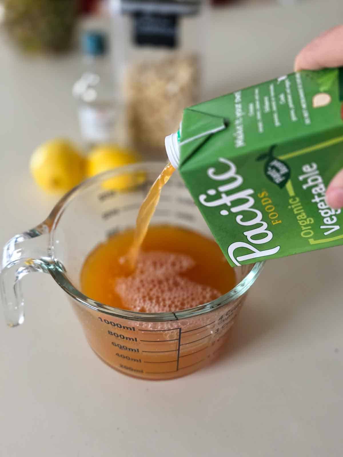 Pouring vegetable broth into a glass measuring cup on a kitchen counter, with lemons and pantry items in the background.