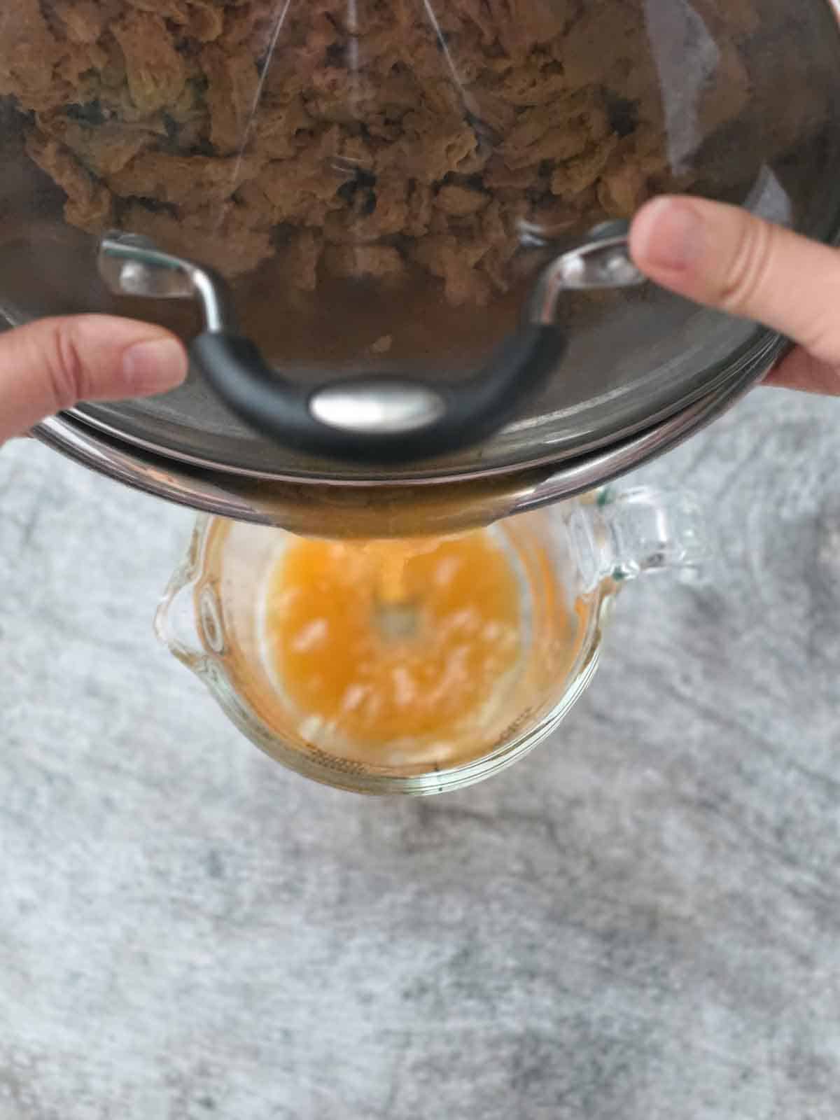Hands holding a lidded bowl and draining excess broth into a measuring cup using her no-squeeze trick.
