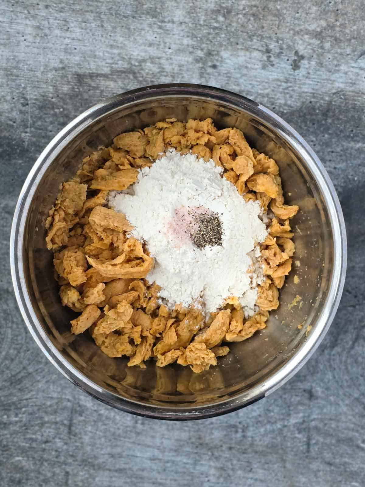Soy curls in a mixing bowl topped with flour, salt, and pepper before tossing.