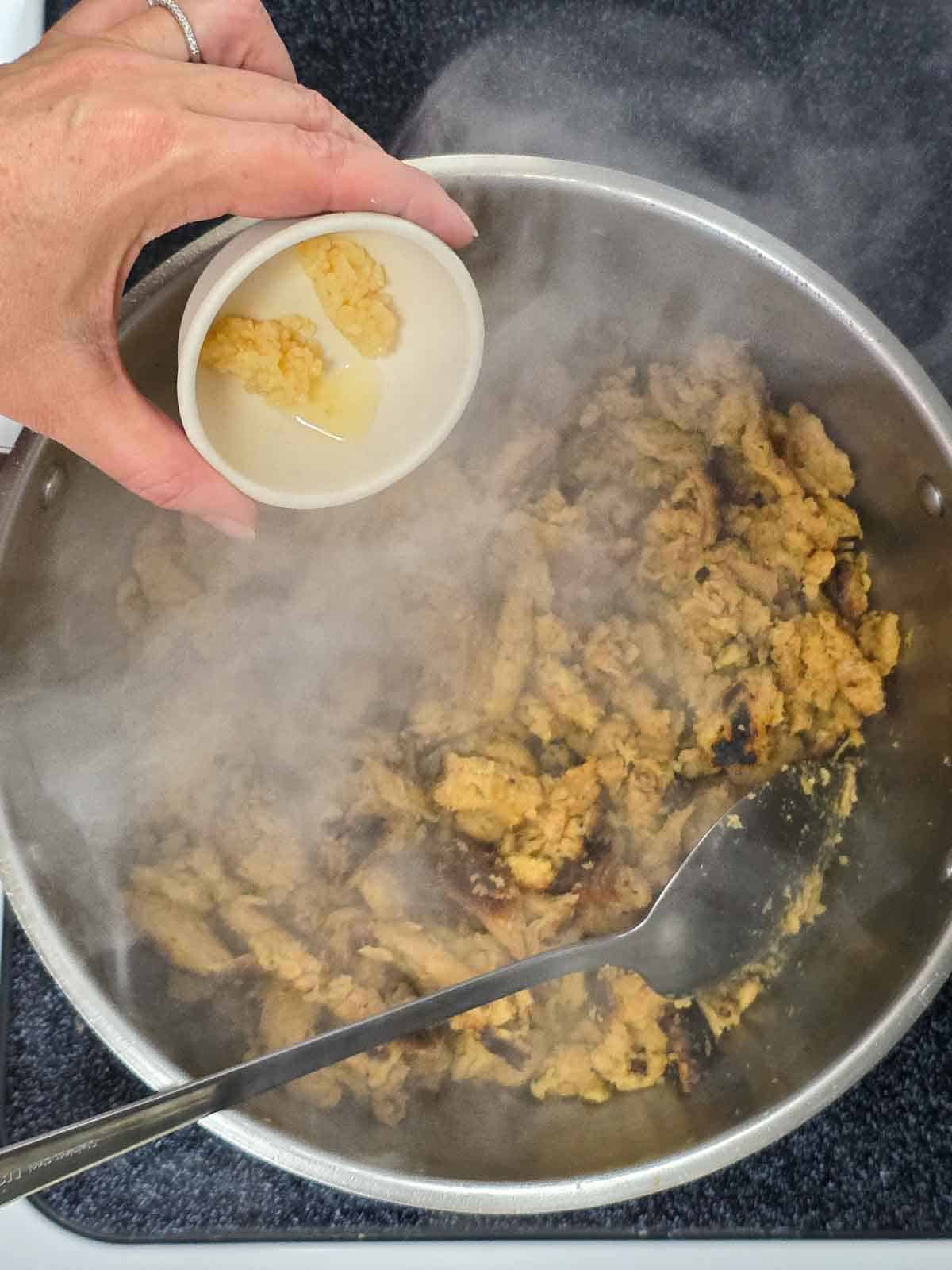 Rhonda adding minced garlic from a small dish into the steaming skillet of soy curls.