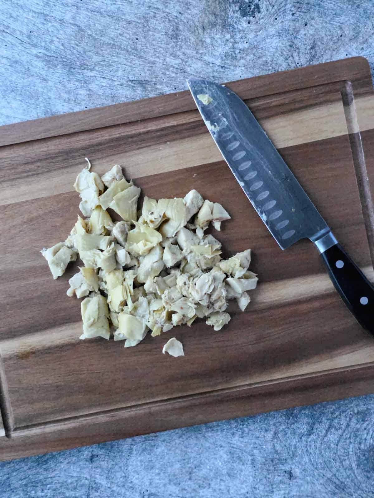 Chopped marinated artichoke hearts on a cutting board next to a chef’s knife.