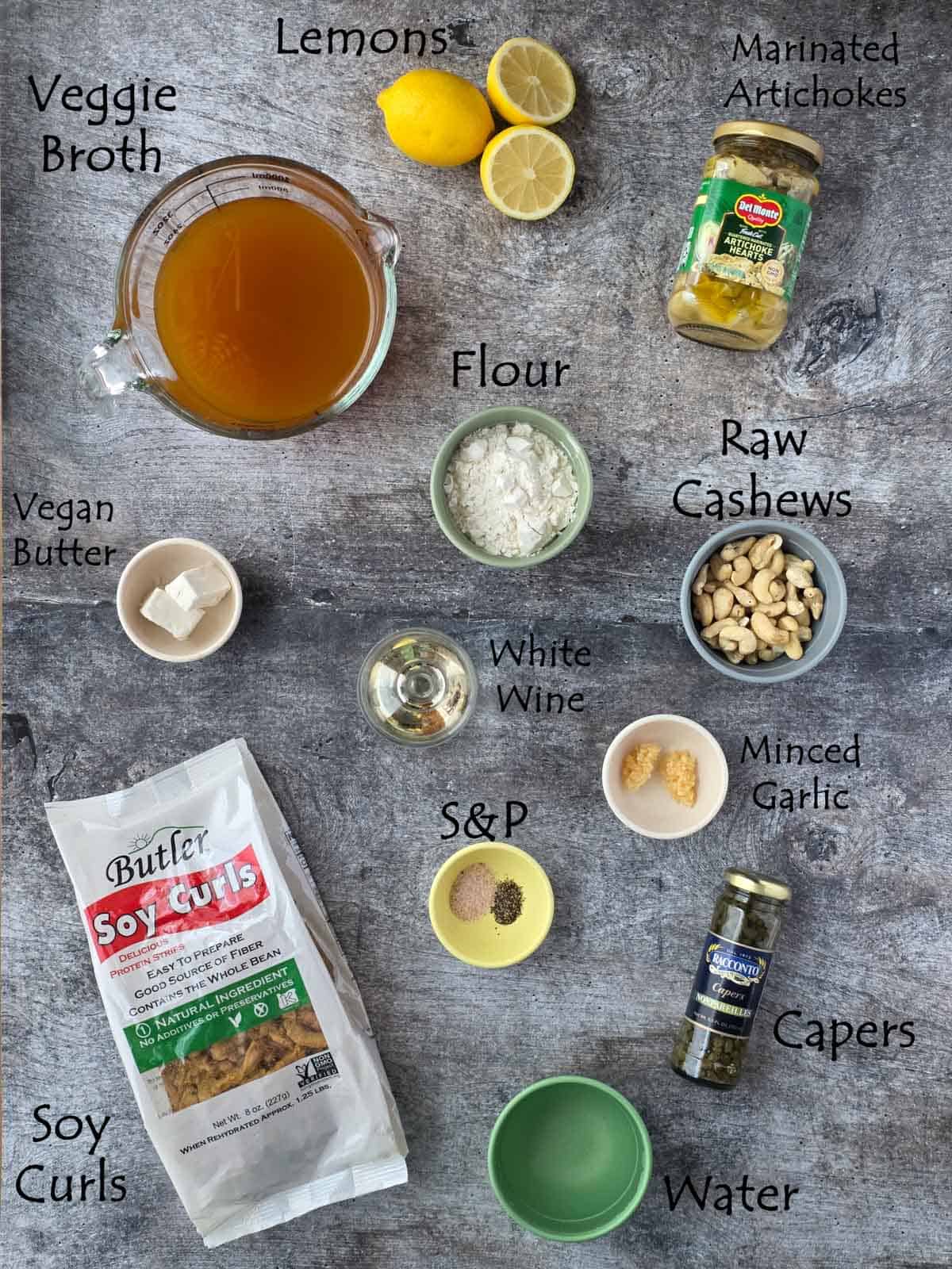 Overhead vegan piccata ingredient flat lay with labels identifying broth, lemons, artichokes, flour, cashews, garlic, soy curls, seasonings, and capers.