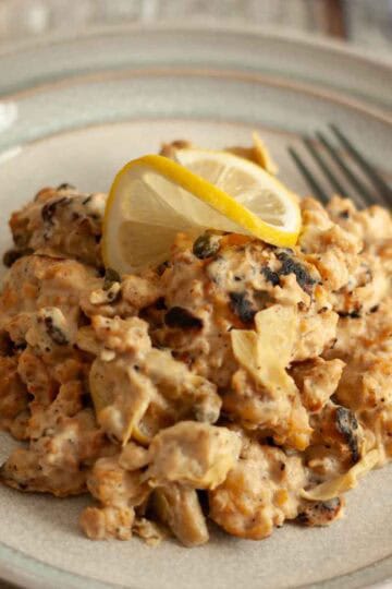 Close-up plate of creamy soy curls with artichokes and capers, topped with a thin lemon slice.