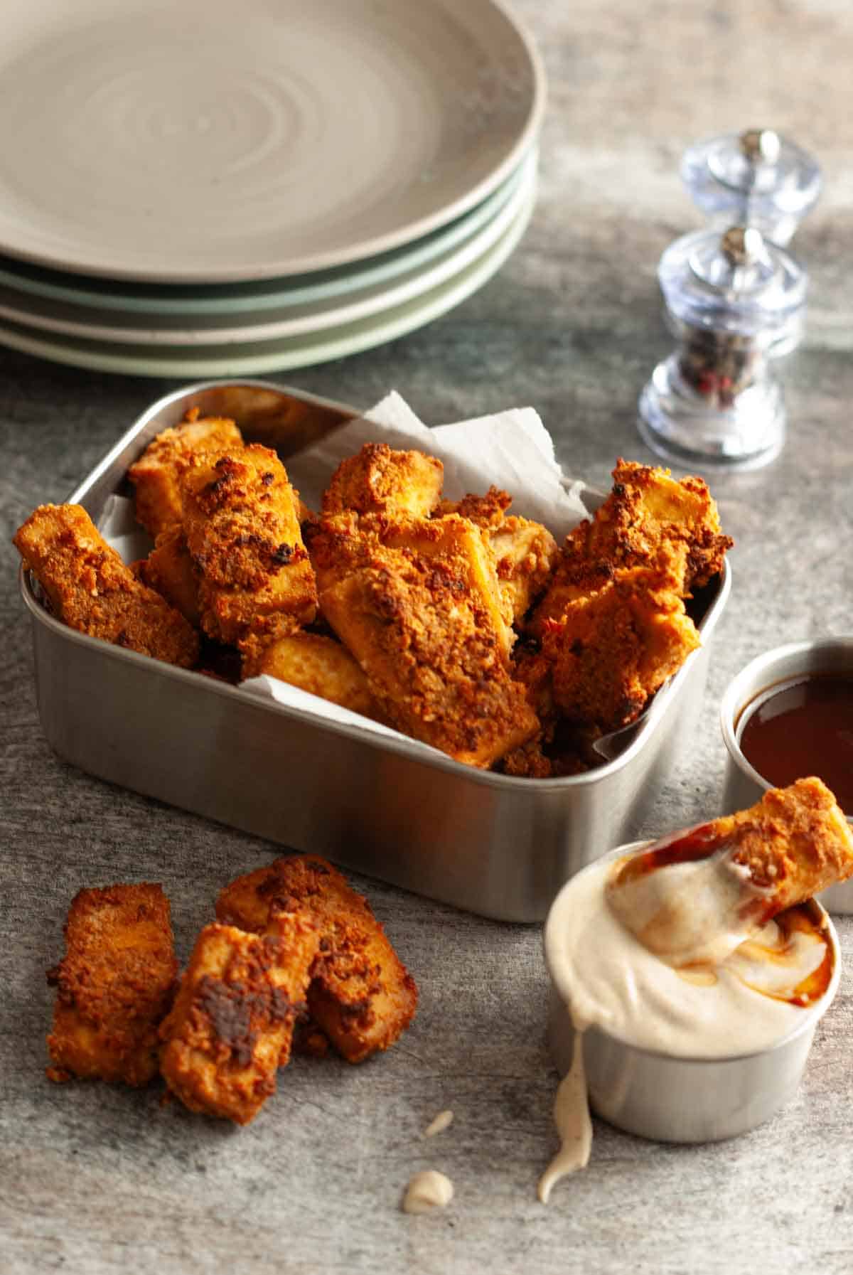 Crispy baked tofu nuggets piled in a metal tray with golden, crunchy edges and a creamy dipping sauce on the side.