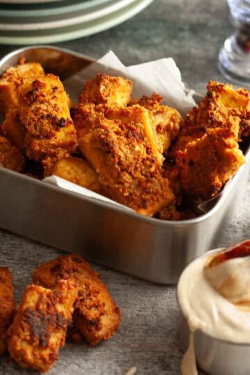 Close-up of crispy baked tofu nuggets with deeply golden coating, served with a creamy sauce for dipping.