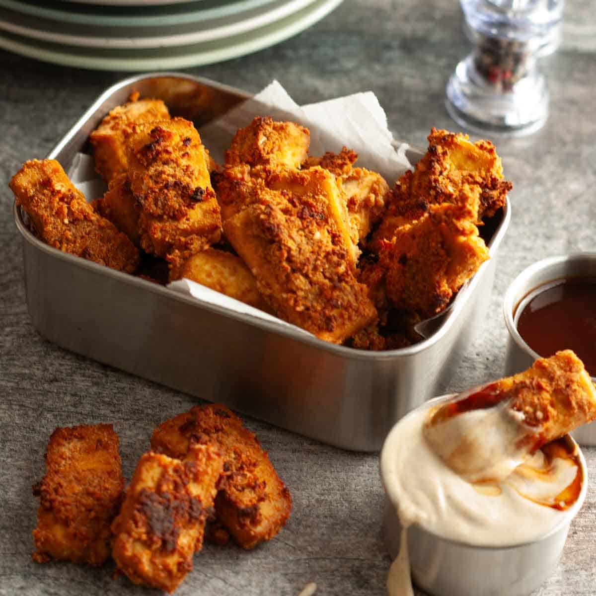 Close-up of crispy baked tofu nuggets with deeply golden coating, served with a creamy sauce for dipping.
