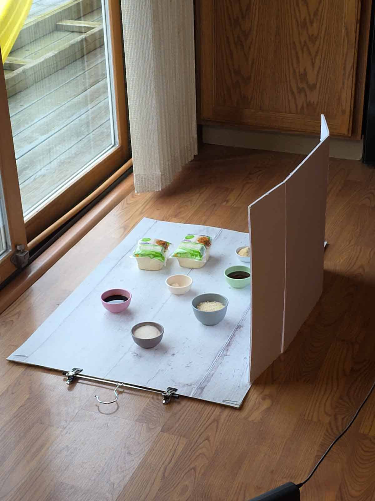 DIY food photography setup on the floor near a window with ingredients arranged on a white backdrop.