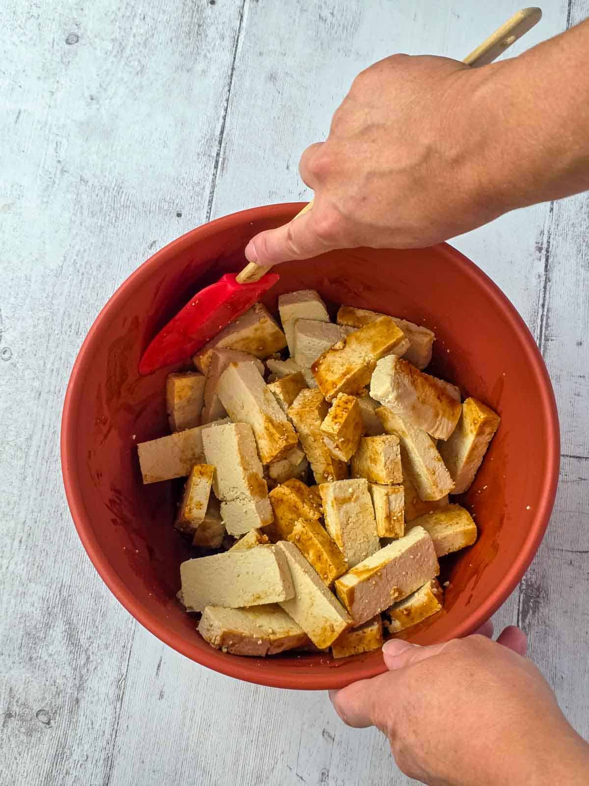Spatula gently folding tofu pieces in seasoning and sauce for making crispy baked tofu nuggets.