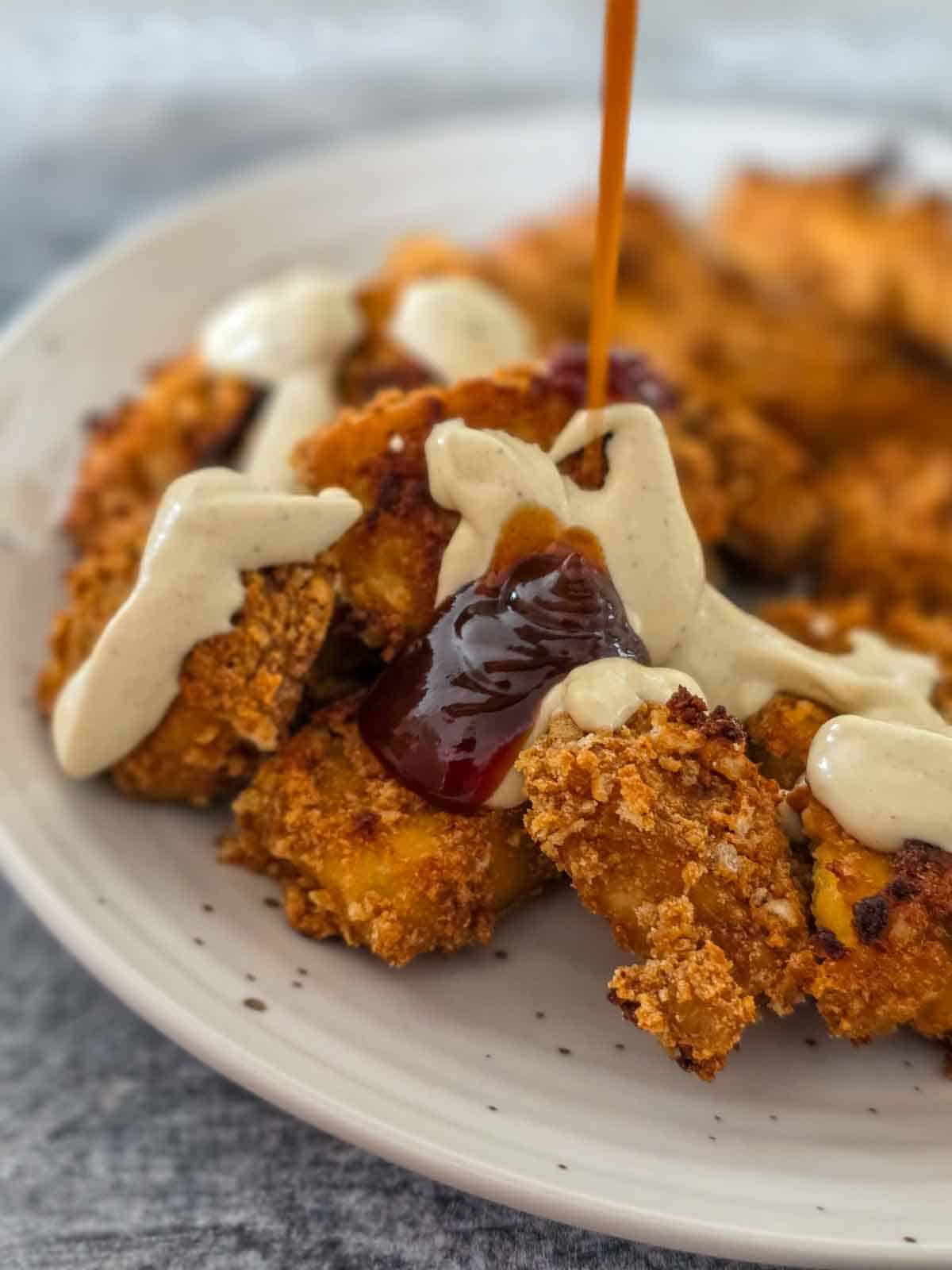 Baked tofu nuggets covered in creamy ranch being drizzled with bbq sauce on a decorative plate.
