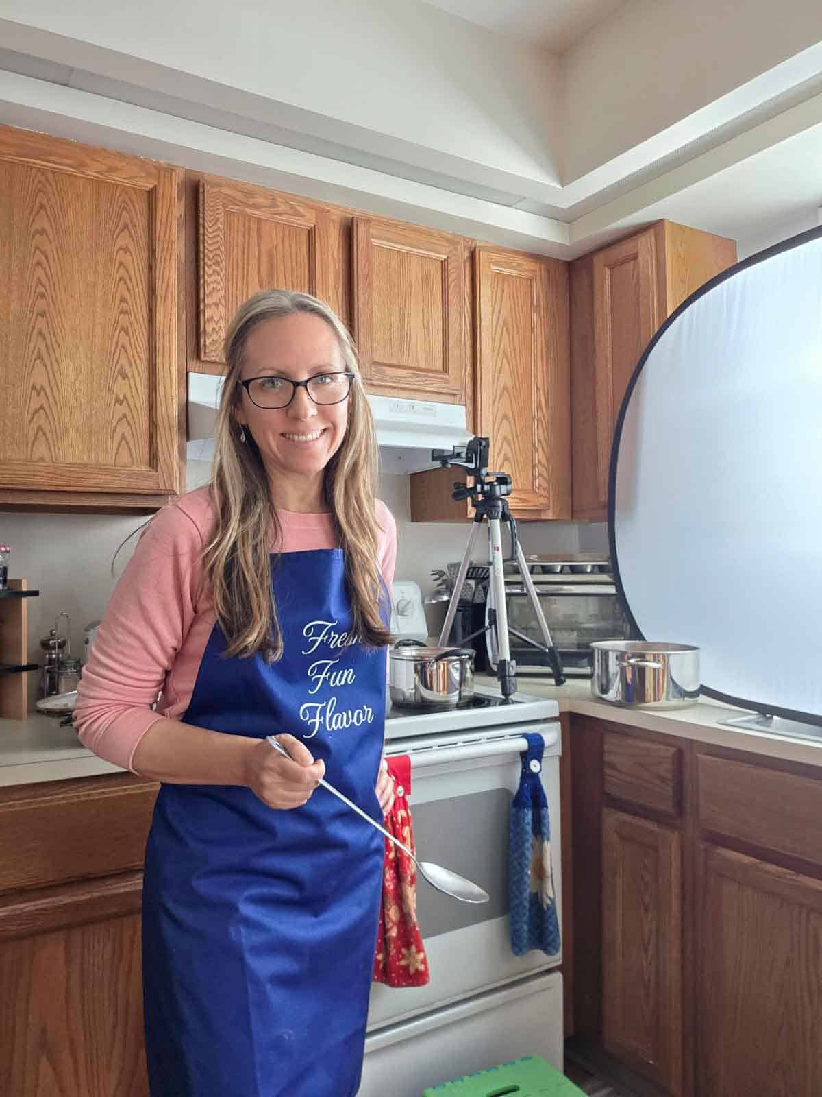 The cook standing in the kitchen wearing an apron, holding a spoon beside the soup pot during the photo shoot.