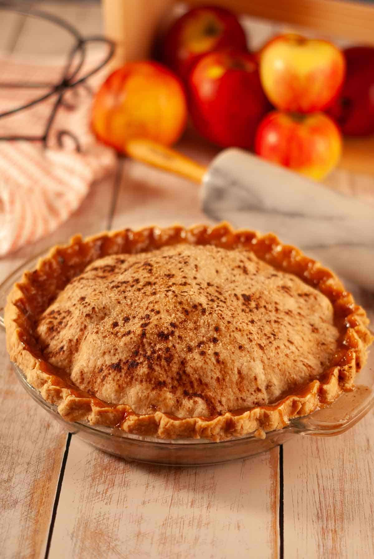 Whole vegan apple pie with golden flaky crust and cinnamon sugar sprinkled on top in a glass pie dish.