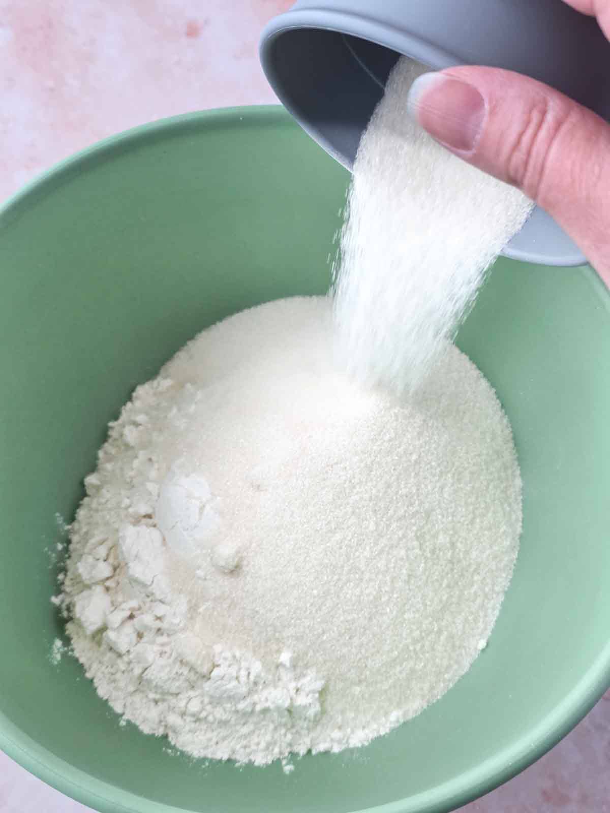 Granulated sugar being poured into a bowl with flour to start the cupcake batter.