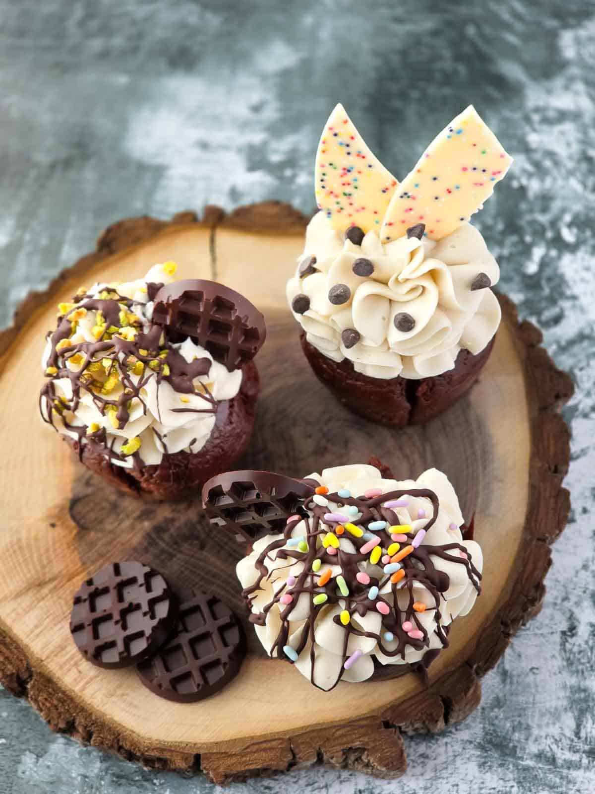 Three frosted chocolate cupcakes with chocolate drizzle and sprinkles on a rustic board.