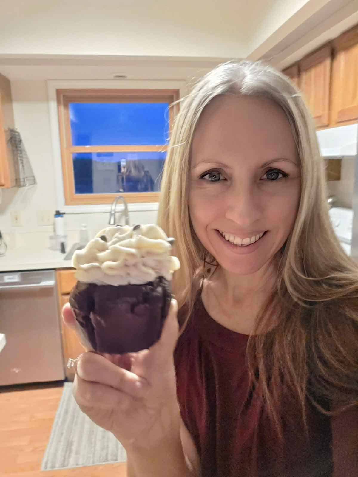 Food blogger smiling and holding a frosted vegan chocolate cupcake in her kitchen.