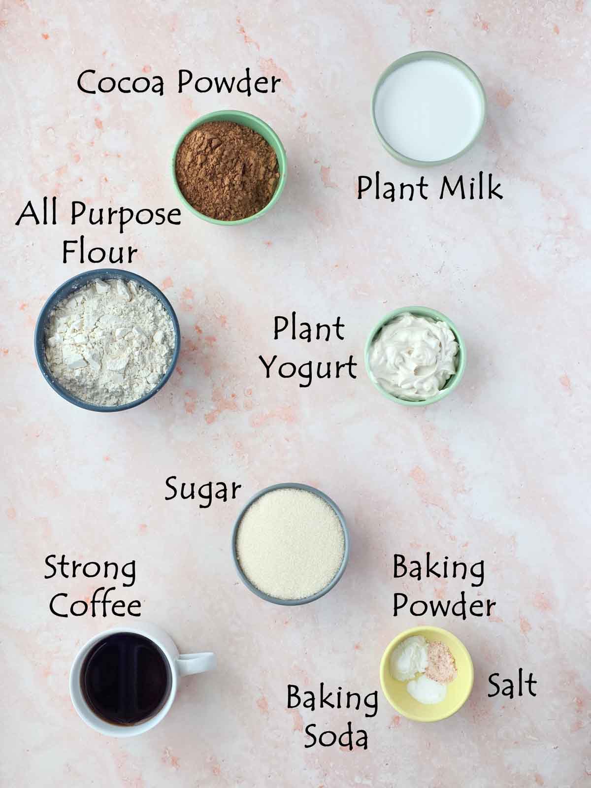 Overhead photo of ingredients for one bowl vegan chocolate cupcakes including flour, cocoa powder, sugar, plant milk, yogurt, coffee, and baking powder.