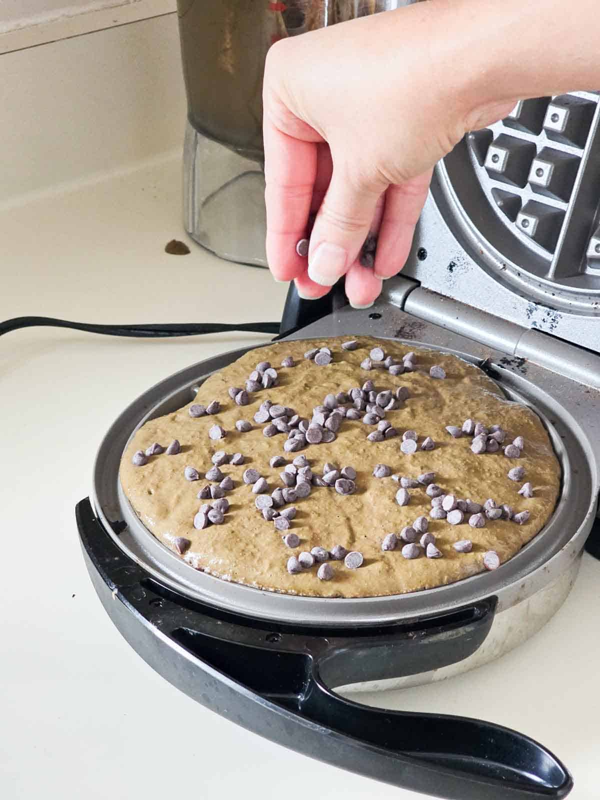 A hand sprinkling chocolate chips onto the waffle batter in the waffle maker.