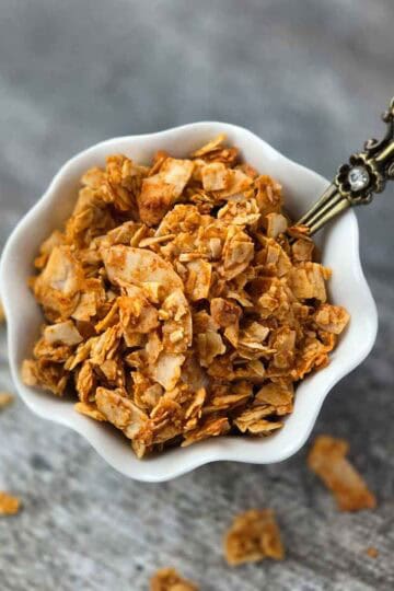Crispy coconut bacon flakes in a small white bowl with a spoon, showing golden brown texture.