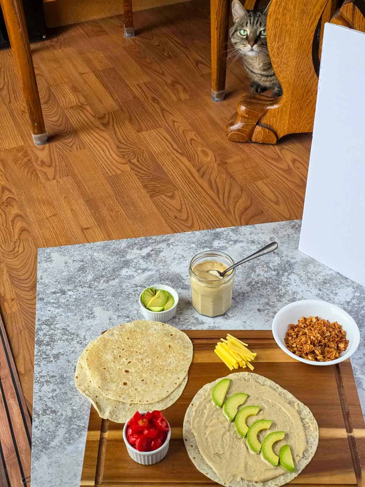 A tabby cat peeking from behind a table at the ingredient flat lay being shot on the floor.