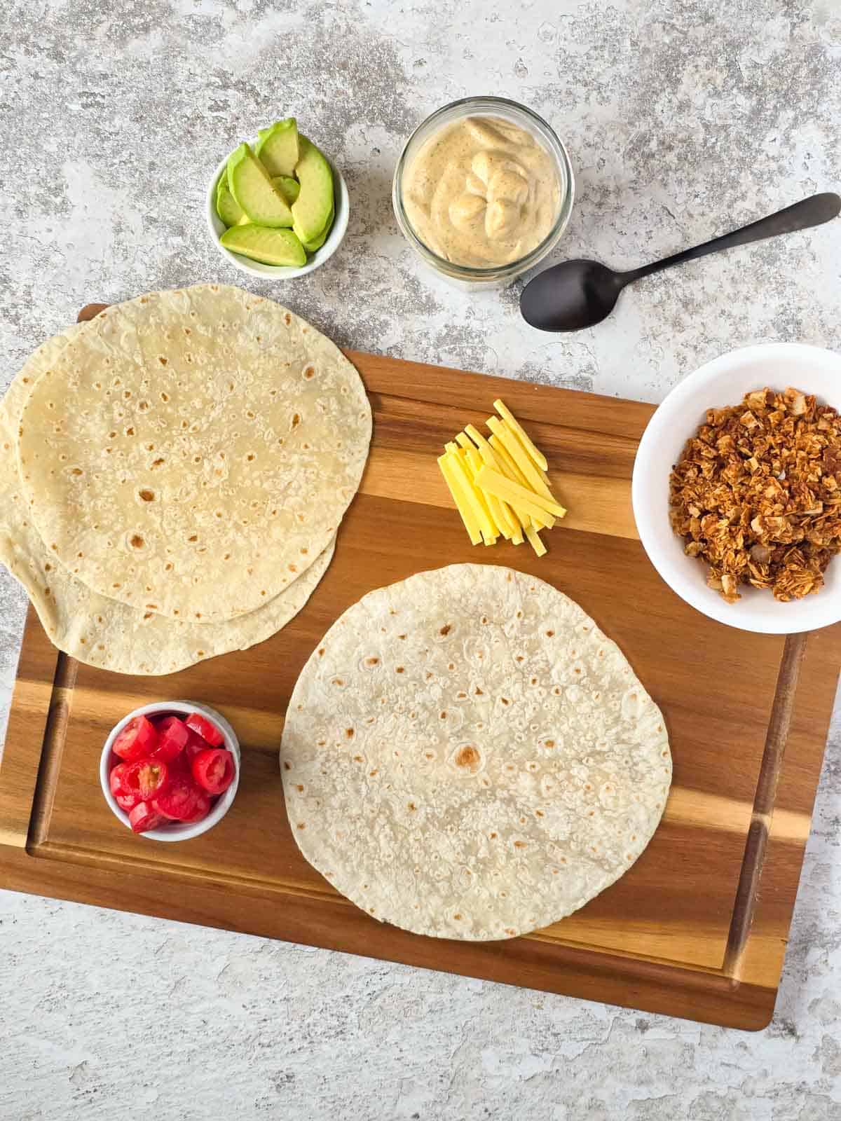 Ingredients needed for a vegan bacon avocado wrap arranged on a cutting board with tortillas, ranch, and toppings.