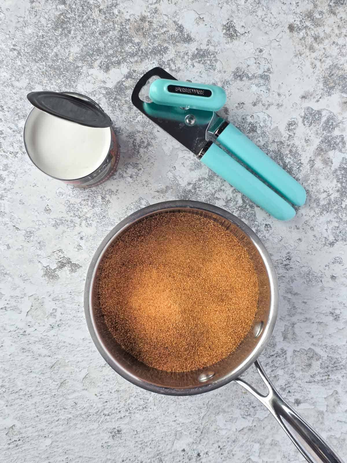 An opened can of coconut milk with a light blue can opener nearby next to a pan filled with coconut sugar.