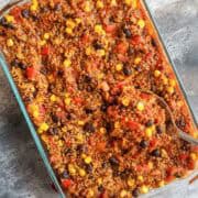 Vegan enchilada bake with corn, black beans, tomatoes, and enchilada sauce being scooped up by a large serving spoon.
