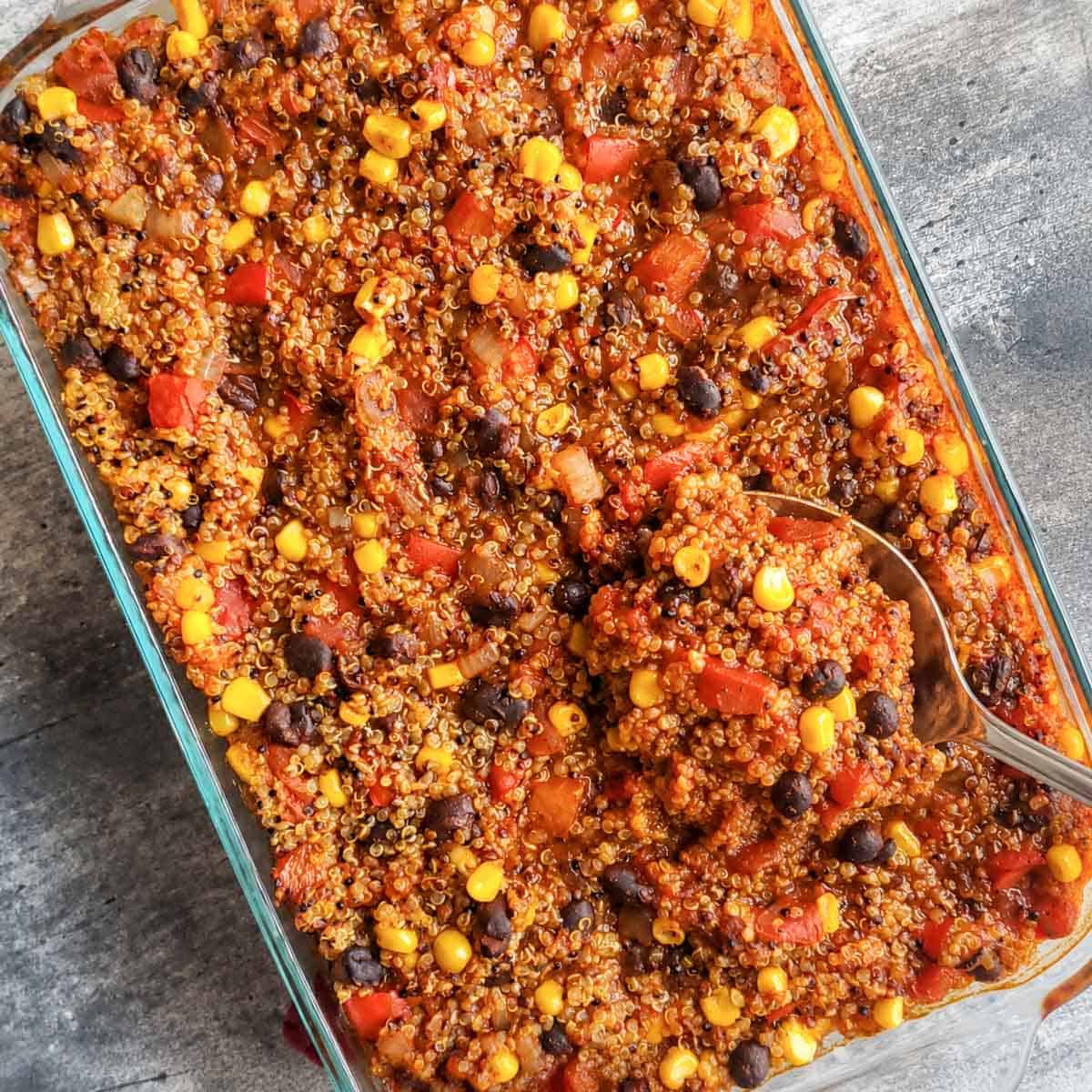 Vegan enchilada bake with corn, black beans, tomatoes, and enchilada sauce being scooped up by a large serving spoon.