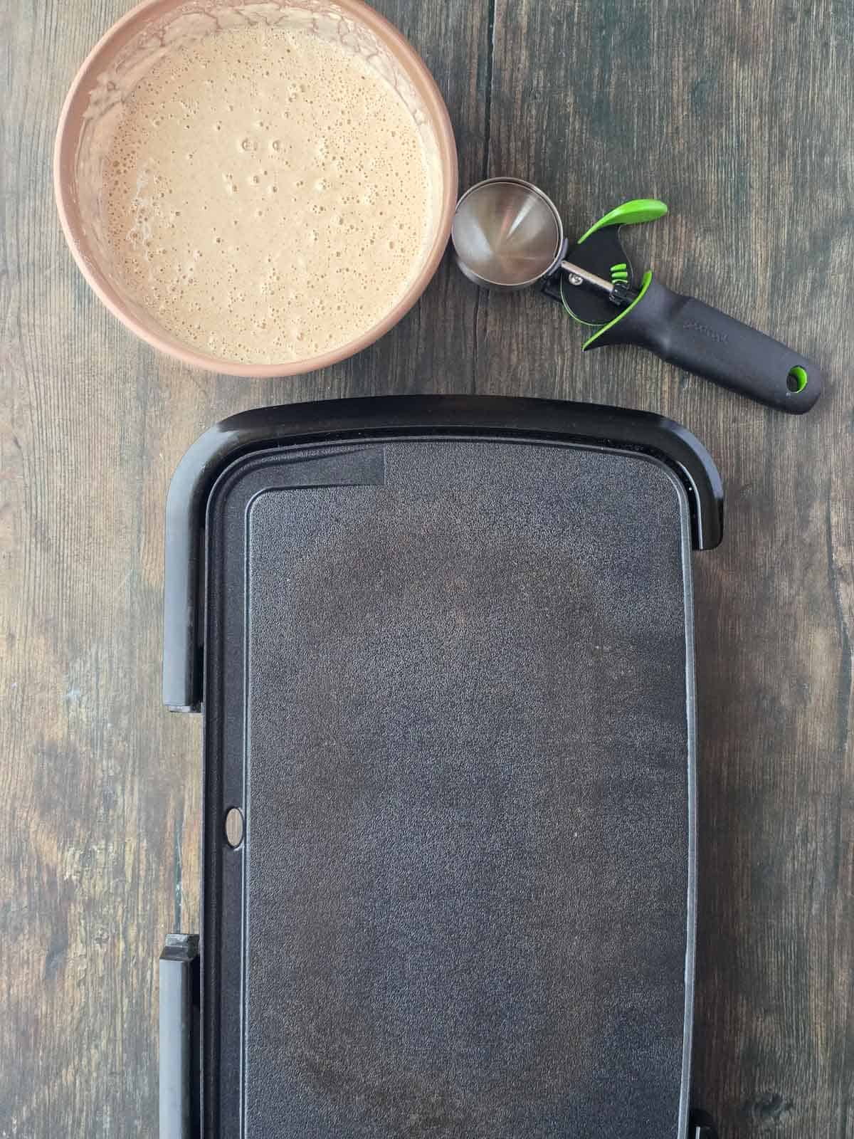 Bowl of prepared pancake batter next to a griddle and portion scoop.