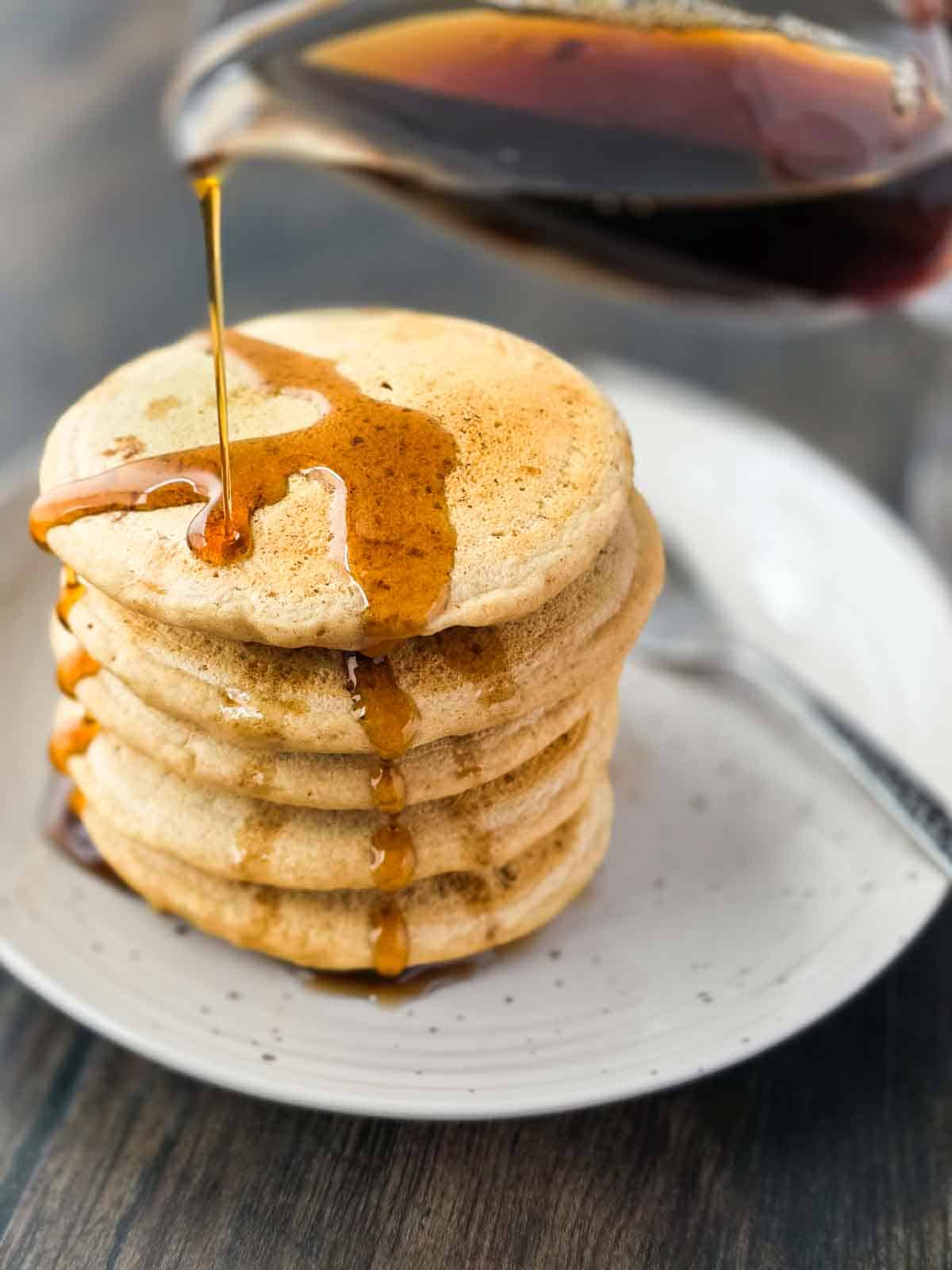 Stack of homemade thick and fluffy pancakes with maple syrup being poured on top and drizzling down the sides.